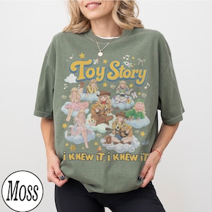 Może przedstawiać: Oliwkowozielony t-shirt z kr&oacute;tkim rękawem z nadrukiem graficznym zawierającym tekst Toy Story oraz i KNEW IT, i KNEW IT. Wz&oacute;r przedstawia wiele postaci w stylu anime siedzących na chmurach, otoczonych gwiazdami, nutami i kokardkami.