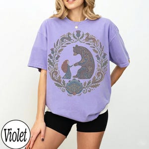 以下が含まれることがあります： バイオレットカラーのTシャツ。円形のデザインで、クマと手をつないでいる人物が描かれています。周りには花柄や装飾的な要素があります。Tシャツは半袖で、ゆったりとしたフィット感です。
