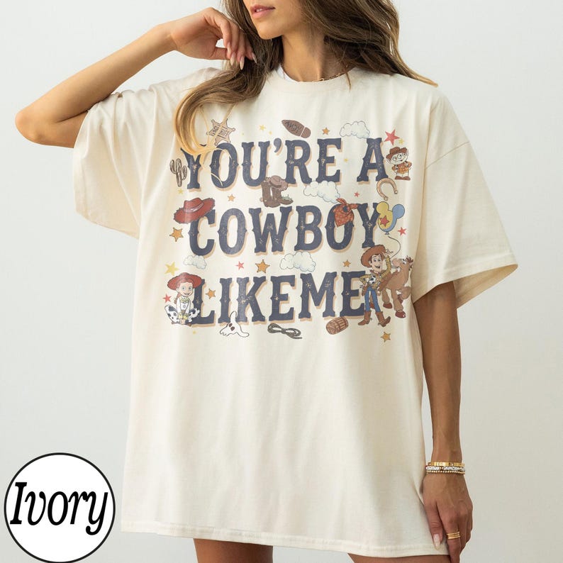 May include: An ivory short-sleeved t-shirt featuring the dark blue text "YOU'RE A COWBOY LIKEME" surrounded by various western-themed illustrations including cowboy hats, boots, stars, clouds, a lasso, and cartoon characters in a vintage style.