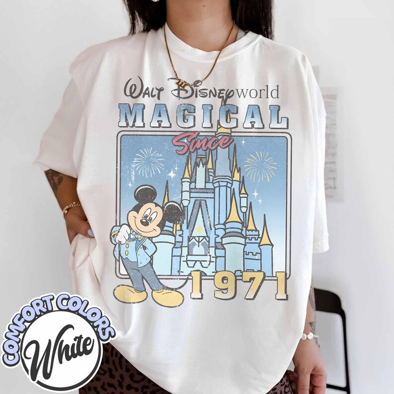May include: White t-shirt featuring a vintage-style graphic of Mickey Mouse in front of Cinderella's castle, with the text "Walt Disney World Magical Since 1971." The shirt also has the words "Comfort Colors" and "White" printed on it.