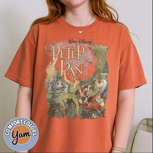 May include: Orange Comfort Colors t-shirt featuring a graphic of Peter Pan. The design includes the text "Walt Disney Peter Pan" in a vintage style, with illustrations of characters from the movie. The shirt has a round neck and short sleeves.