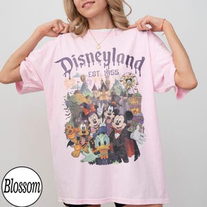 Puede incluir: Una camiseta rosa claro con un estampado gr&aacute;fico de personajes de Disneyland con disfraces de Halloween, incluyendo a Mickey Mouse, Minnie Mouse, Goofy, el Pato Donald y Pluto. El texto dice Disneyland EST. 1955 sobre un castillo.