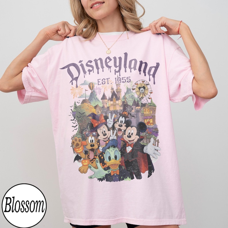 Puede incluir: Una camiseta rosa claro con un estampado gr&aacute;fico de personajes de Disneyland con disfraces de Halloween, incluyendo a Mickey Mouse, Minnie Mouse, Goofy, el Pato Donald y Pluto. El texto dice Disneyland EST. 1955 sobre un castillo.