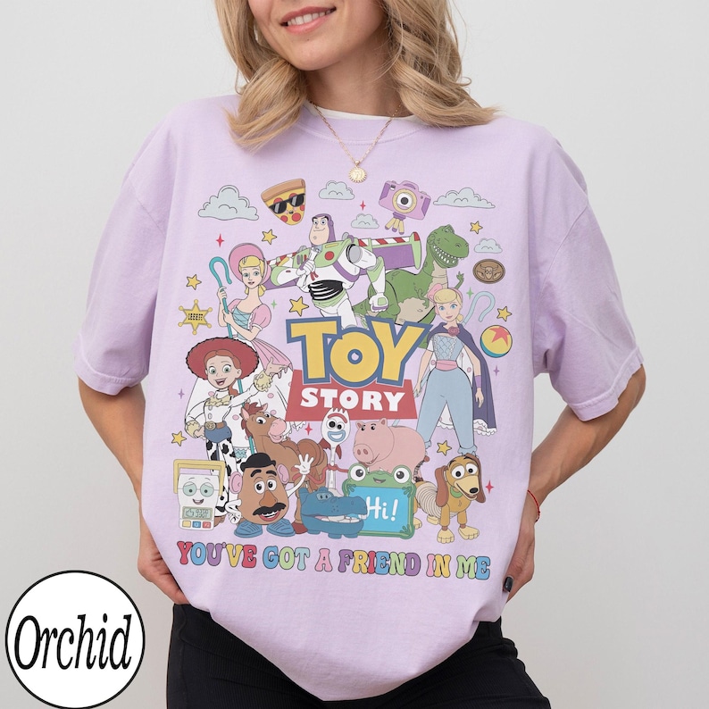 May include: A light purple t-shirt featuring a colorful graphic print of various Toy Story characters including Buzz Lightyear, Woody, Jessie, and Rex. The text Toy Story and You've Got A Friend In Me is printed in bold, multi-colored lettering.