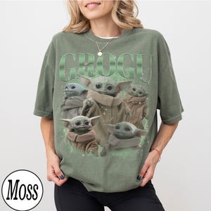 Puede incluir: Camiseta verde con la palabra "GROGU" en una fuente degradada verde sobre un gr&aacute;fico de Grogu de The Mandalorian. El gr&aacute;fico muestra a Grogu en varias poses. Un c&iacute;rculo blanco con la palabra "Moss" est&aacute; en la esquina inferior izquierda.