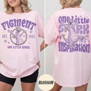May include: A light pink t-shirt featuring a purple dragon graphic with the text "Figment" and "One Little Spark" on the front. The back of the shirt has the text "One Little Spark of Inspiration" and the same dragon graphic.