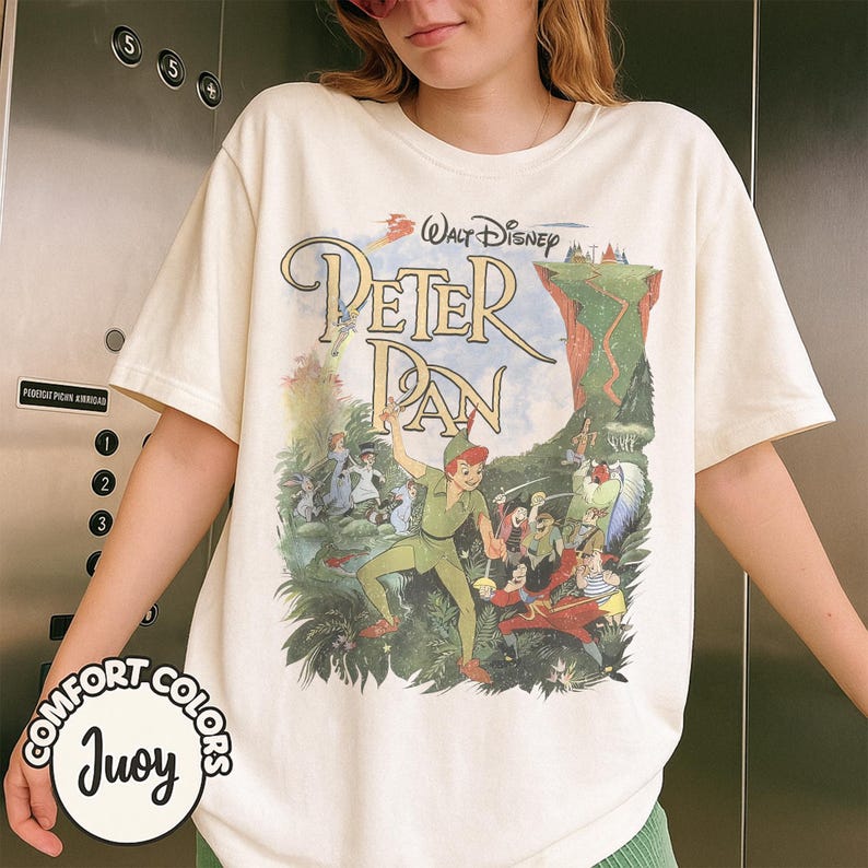 May include: Cream-colored t-shirt featuring a colorful graphic of Peter Pan from Walt Disney. The design includes the title "Peter Pan" in a whimsical font, along with illustrations of characters and scenes from the movie. The shirt has short sleeves and a relaxed fit.