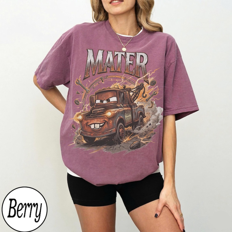 May include: Berry-colored t-shirt featuring a graphic of Mater from the Cars movie. The design includes the word "MATER" above the character, with lightning bolts and tools surrounding the image. The shirt has a relaxed fit.
