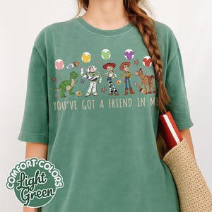 May include: Light green Comfort Colors t-shirt featuring a graphic of characters from the movie Toy Story. The text "YOU'VE GOT A FRIEND IN ME" is printed below the characters. The shirt has short sleeves and a crew neck.