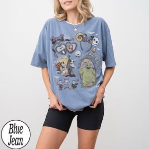 May include: A blue jean t-shirt featuring a colorful graphic of characters from The Nightmare Before Christmas. The design includes Jack Skellington, Sally, Oogie Boogie, and other iconic elements from the movie. The shirt has a round neckline and short sleeves.