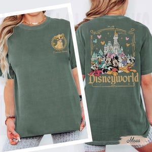 May include: Olive green t-shirt featuring a Simba graphic on the front and a Disneyworld graphic on the back. The Disneyworld graphic includes Mickey Mouse, Minnie Mouse, Donald Duck, Daisy Duck, and Goofy. The shirt has the text "Disneyworld 1971".