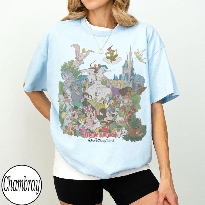 May include: Light blue t-shirt featuring a colorful graphic of Disney characters, including Mickey Mouse, Dumbo, and Peter Pan, with the text "Magic Kingdom" and "Walt Disney World". The shirt has short sleeves and a crew neck.