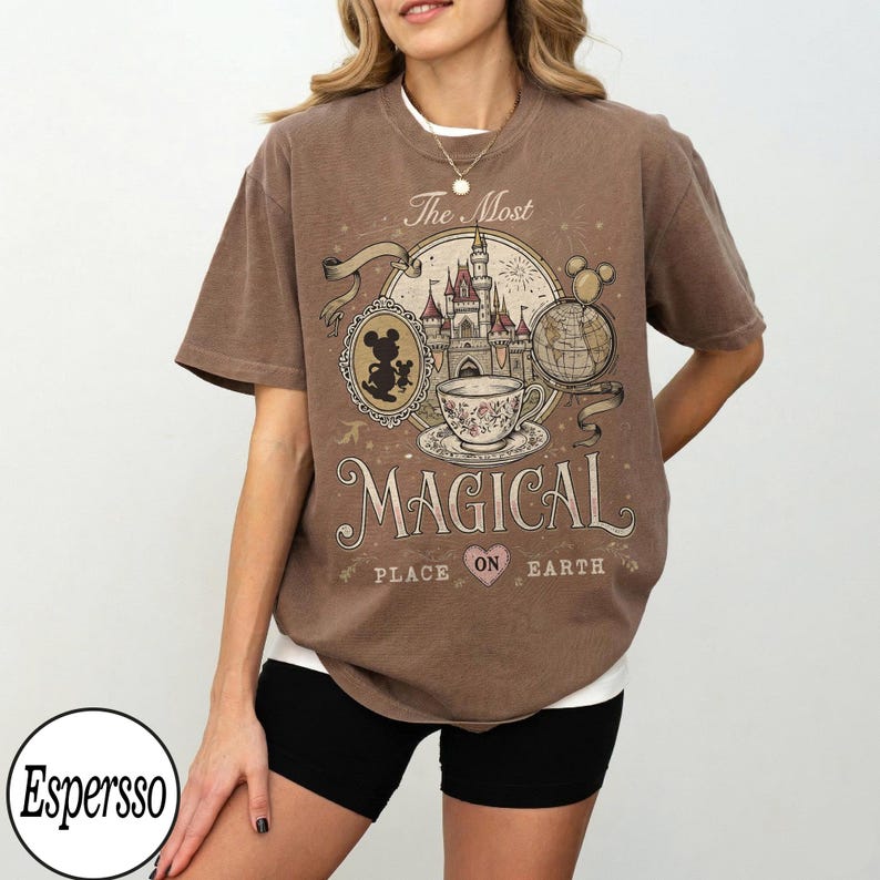 May include: Brown t-shirt with a vintage-style graphic featuring a castle, teacup, Mickey Mouse silhouette, and the words "The Most Magical Place on Earth." The design includes a globe and fireworks. The shirt has short sleeves.