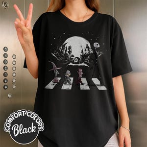 Peut inclure: T-shirt noir Comfort Colors avec un motif de personnages traversant une rue, avec une pleine lune et des b&acirc;timents effrayants en arri&egrave;re-plan. Le motif comprend Jack Skellington et d'autres personnages de L'&Eacute;trange No&euml;l de Monsieur Jack.