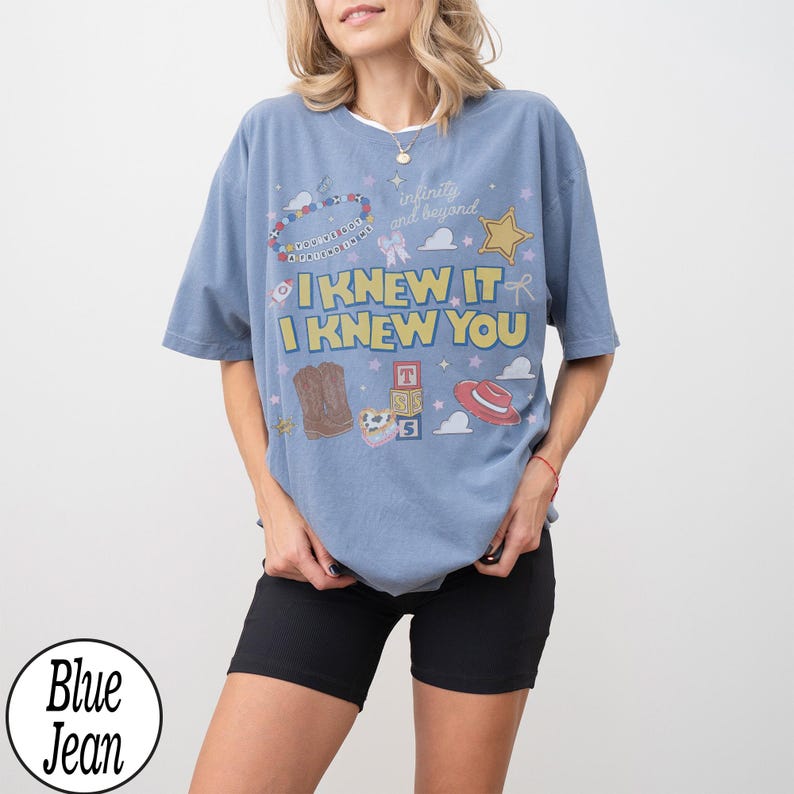 May include: A blue oversized t-shirt featuring a graphic print with the text I KNEW IT I KNEW YOU, along with illustrations of a cowboy hat, boots, alphabet blocks, a star, and a beaded bracelet with the text YOU'VE GOT A FRIEND IN ME.