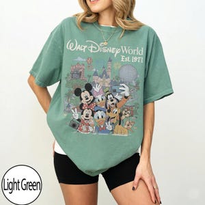 May include: A light green short-sleeve t-shirt featuring a graphic print of Walt Disney World Est. 1971. The design includes Mickey Mouse, Minnie Mouse, Donald Duck, Daisy Duck, Goofy, Pluto, and Chip and Dale taking a selfie in front of the park.