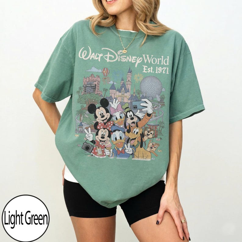 May include: A light green short-sleeve t-shirt featuring a graphic print of Walt Disney World Est. 1971. The design includes Mickey Mouse, Minnie Mouse, Donald Duck, Daisy Duck, Goofy, Pluto, and Chip and Dale taking a selfie in front of the park.