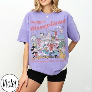 May include: A lilac-coloured t-shirt with a Disneyland graphic featuring Mickey Mouse, Donald Duck, and other characters. The text "CALIFORNIA Est. 1955 Disneyland" is printed on the front. The shirt has a relaxed fit.