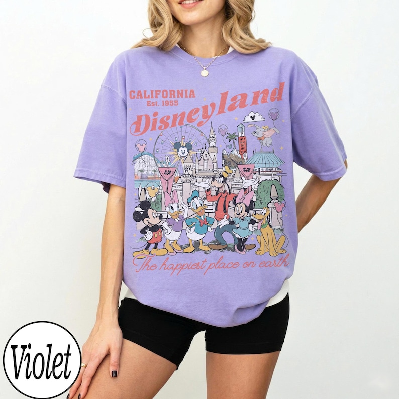 May include: A lilac-coloured t-shirt with a Disneyland graphic featuring Mickey Mouse, Donald Duck, and other characters. The text "CALIFORNIA Est. 1955 Disneyland" is printed on the front. The shirt has a relaxed fit.