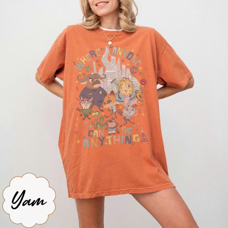 May include: Orange t-shirt with a cartoon graphic and the words "WHERE ANYONE CAN BE ANYTHING". The design includes a castle and animal characters. The shirt has short sleeves and a relaxed fit.