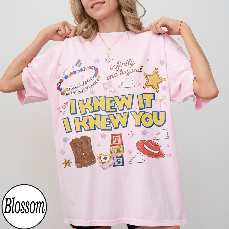 May include: A light pink t-shirt featuring a graphic print with the text "I KNEW IT I KNEW YOU" and "infinity and beyond". The design includes illustrations of a beaded bracelet, a gold star, cowboy boots, alphabet blocks, and a red hat.