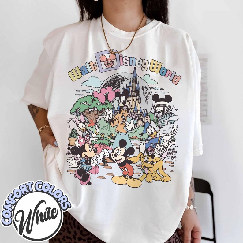 May include: White Comfort Colors t-shirt featuring a colorful graphic of Disney characters and the text "Walt Disney World". The design includes Mickey Mouse, Minnie Mouse, Donald Duck, and other characters in front of a castle.