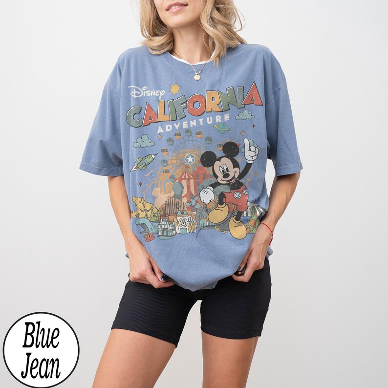 May include: A blue t-shirt with a graphic of Mickey Mouse and the words "Disney California Adventure". The design includes a Ferris wheel, a rocket ship, and other amusement park elements. The shirt is a light blue color.