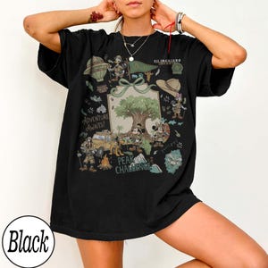 May include: Black t-shirt with a vibrant graphic. The design includes cartoon characters, a tree, and the text "Adventure Awaits!" and "Peak Challenge." The graphic features a jeep, hot air balloon, and safari-themed elements.