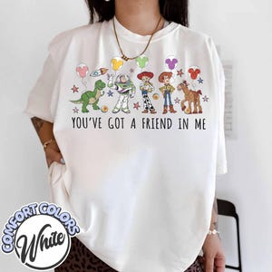 May include: White Comfort Colors t-shirt featuring a colorful graphic of Toy Story characters, including Rex, Buzz Lightyear, Woody, and Jessie. The text "YOU'VE GOT A FRIEND IN ME" is printed below the characters. The shirt has a relaxed fit.