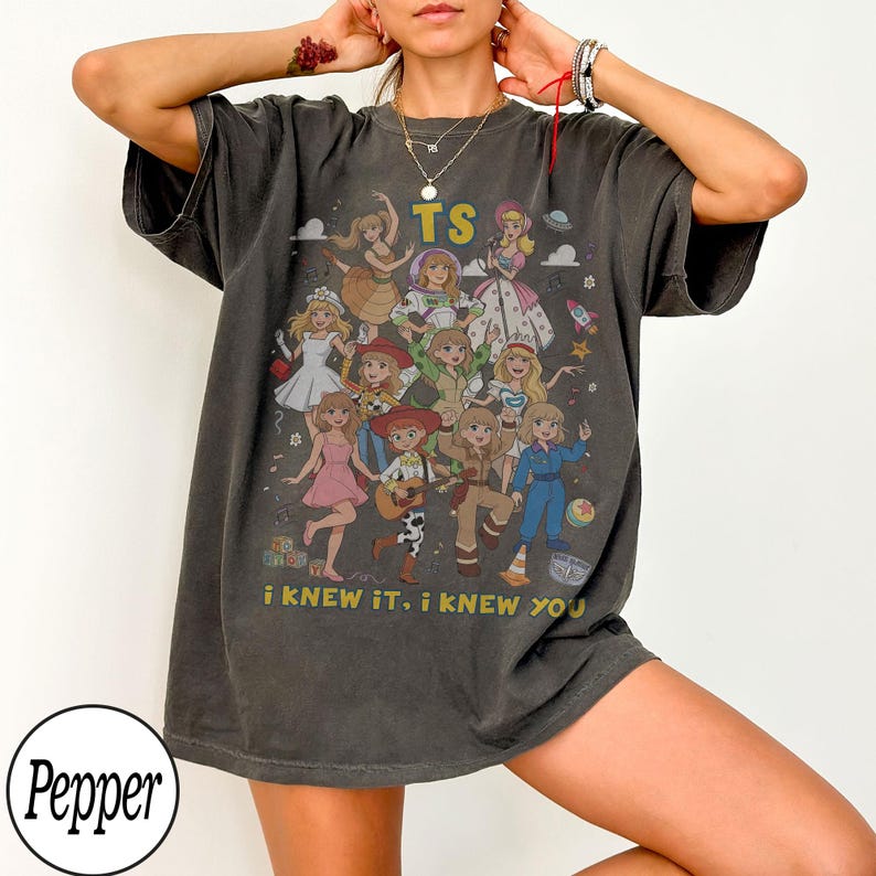 May include: A charcoal gray short-sleeve t-shirt featuring a graphic print of multiple animated characters in various outfits. The text TS is printed at the top, and the text i KNEW IT, i KNEW YOU is printed at the bottom in yellow lettering.