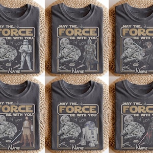 May include: Six gray t-shirts with the phrase "MAY THE FORCE BE WITH YOU" and the word "NAME" printed on them. Each shirt features a different Star Wars character or scene, including the Millennium Falcon, Stormtrooper, and Darth Vader.