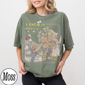 May include: An olive green short-sleeve t-shirt featuring a graphic print of various animated characters and the text "I KNEW IT, I KNEW YOU TS". The design includes a central figure playing a guitar, surrounded by multiple characters, a star-shaped badge, and a ball, all enclosed within a thin rectangular border.