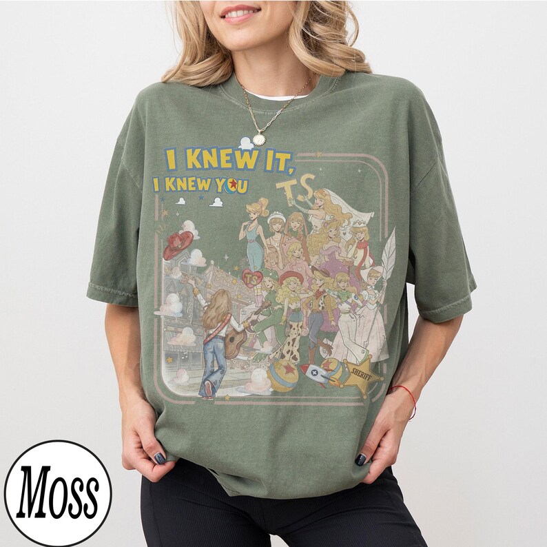 May include: An olive green short-sleeve t-shirt featuring a graphic print of various animated characters and the text "I KNEW IT, I KNEW YOU TS". The design includes a central figure playing a guitar, surrounded by multiple characters, a star-shaped badge, and a ball, all enclosed within a thin rectangular border.