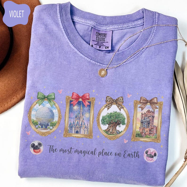 K&ouml;nnte beinhalten: Ein violettes Comfort Colors T-Shirt mit vier gerahmten Illustrationen von Themenpark-Wahrzeichen. Das Shirt tr&auml;gt den Text "The most magical place on Earth" und ist mit einer goldenen Halskette versehen.