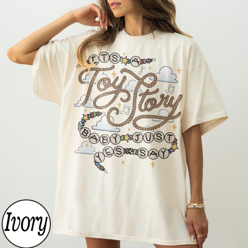 May include: An ivory short-sleeved t-shirt featuring a graphic print with the text "IT'S A Toy Story BABY JUST YES SAY". The design includes brown rope-style lettering, colorful beads, blue clouds, musical notes, and yellow stars on the front.