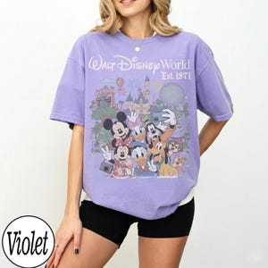 May include: A violet short-sleeve t-shirt featuring a graphic print of Walt Disney World Est. 1971 with various cartoon characters including Mickey Mouse, Minnie Mouse, Donald Duck, Daisy Duck, Goofy, Pluto, and Chip 'n Dale taking a selfie.