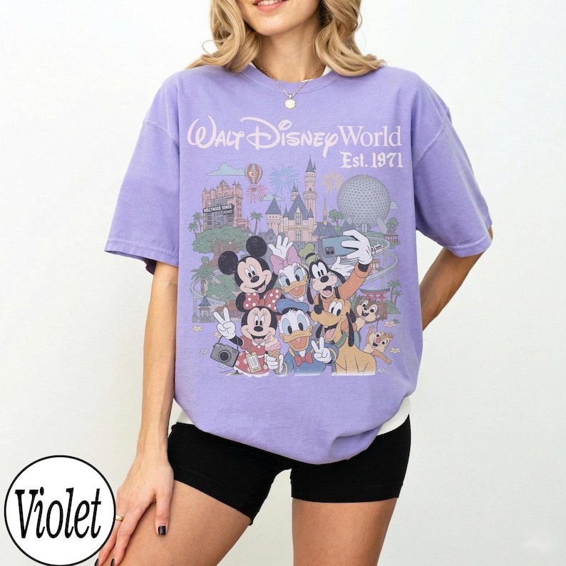 May include: A violet short-sleeve t-shirt featuring a graphic print of Walt Disney World Est. 1971 with various cartoon characters including Mickey Mouse, Minnie Mouse, Donald Duck, Daisy Duck, Goofy, Pluto, and Chip 'n Dale taking a selfie.