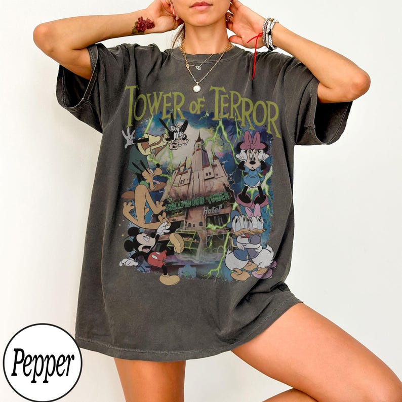 K&ouml;nnte beinhalten: Dunkelgraues T-Shirt mit einer Grafik von Disney-Figuren vor dem Hollywood Tower Hotel, mit dem Text "Tower of Terror" in Gr&uuml;n. Das Design umfasst Micky Maus, Minnie Maus, Donald Duck und Goofy. Das Shirt hat kurze &Auml;rmel.