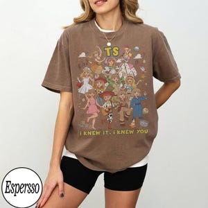 May include: A brown short-sleeve t-shirt featuring a graphic print of multiple cartoon characters in various outfits. The text TS is printed in yellow at the top, and the text i knew it, i knew you is printed in yellow at the bottom of the graphic.