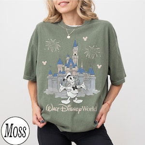 May include: Olive green t-shirt with a graphic of Donald Duck in front of Cinderella's castle at Walt Disney World. The design includes fireworks and Mickey Mouse silhouettes. The text "Walt Disney World" is below the castle.