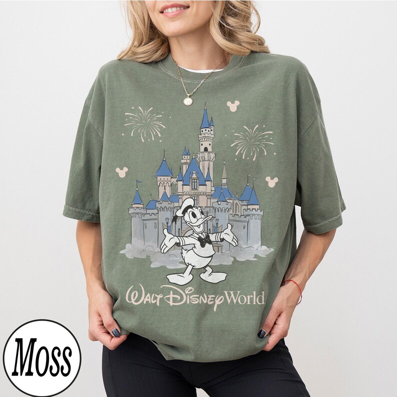 May include: Olive green t-shirt with a graphic of Donald Duck in front of Cinderella's castle at Walt Disney World. The design includes fireworks and Mickey Mouse silhouettes. The text "Walt Disney World" is below the castle.