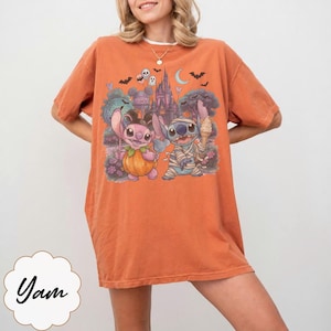 May include: An orange short-sleeve t-shirt featuring a graphic print of two cartoon characters, one dressed as a pumpkin and the other as a mummy, standing in front of a castle with bats, ghosts, and a crescent moon in the background.
