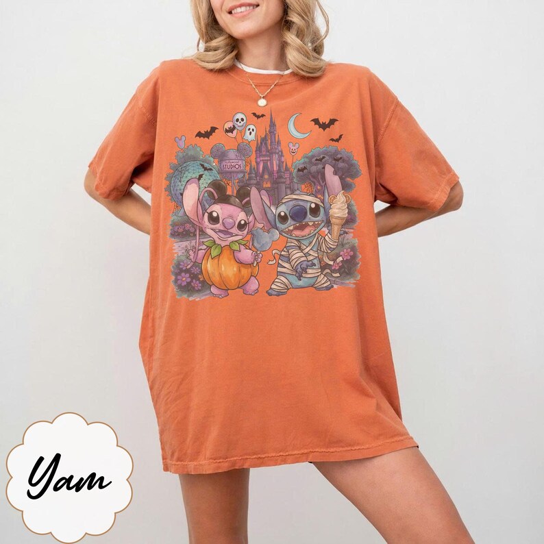 May include: An orange short-sleeve t-shirt featuring a graphic print of two cartoon characters, one dressed as a pumpkin and the other as a mummy, standing in front of a castle with bats, ghosts, and a crescent moon in the background.