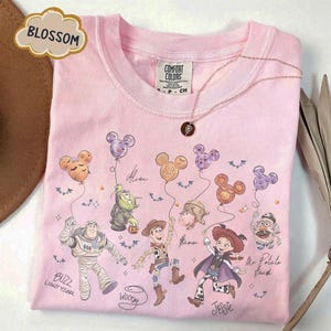 Pode incluir: Camiseta Comfort Colors rosa claro com personagens de Toy Story da Disney. A camiseta de gola redonda apresenta Buzz Lightyear, Woody, Jessie e outros personagens segurando bal&otilde;es em forma de Mickey Mouse. A palavra "Blossom" est&aacute; em uma caixa de texto em forma de nuvem.