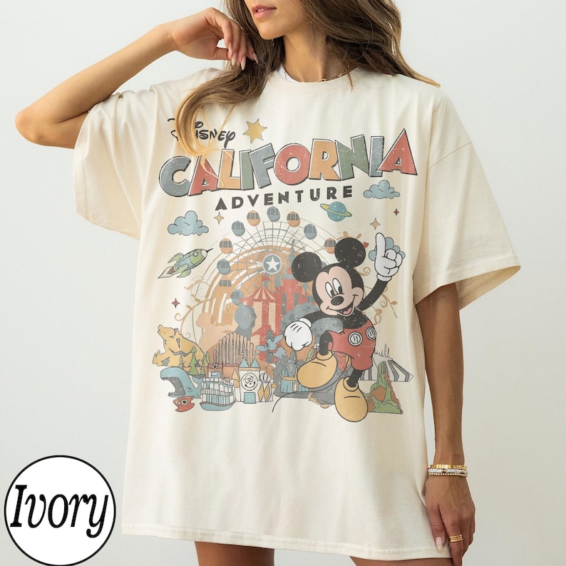 May include: Ivory-colored t-shirt featuring a graphic of Mickey Mouse and the words "Disney California Adventure." The design includes a Ferris wheel, clouds, and a rocket ship. The shirt has short sleeves and a relaxed fit.