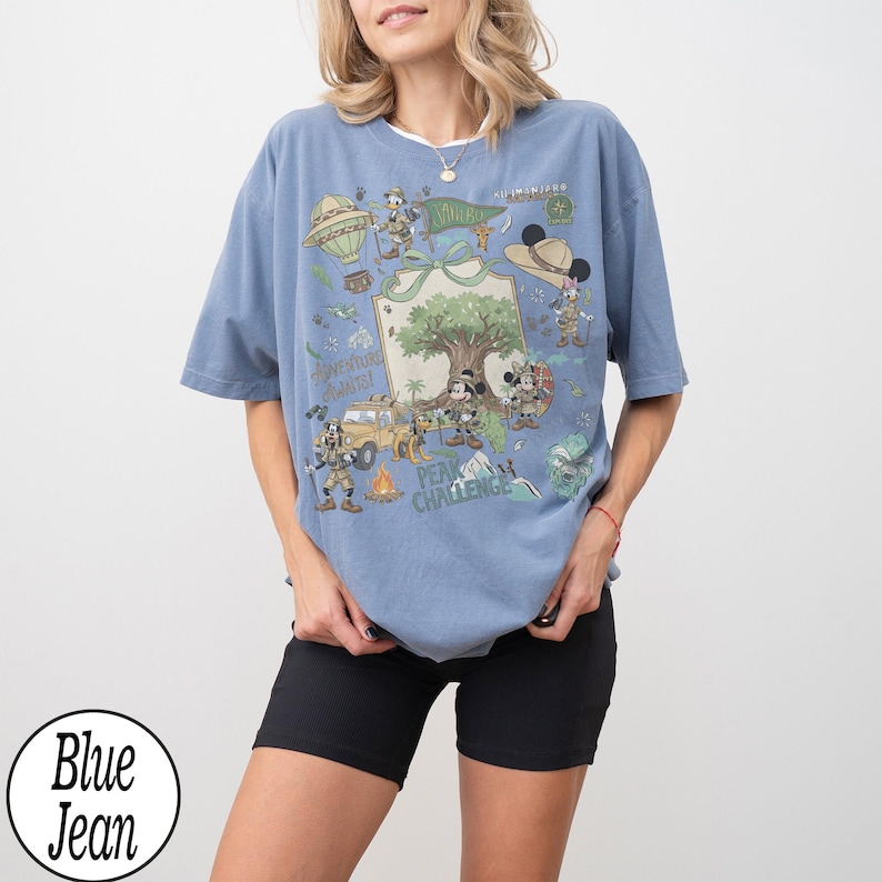 May include: A blue t-shirt with a graphic featuring cartoon characters, a tree, and the text "Adventure Awaits! Peak Challenge." The design includes a hot air balloon, a jeep, and other adventure-themed elements. The shirt is a relaxed fit.