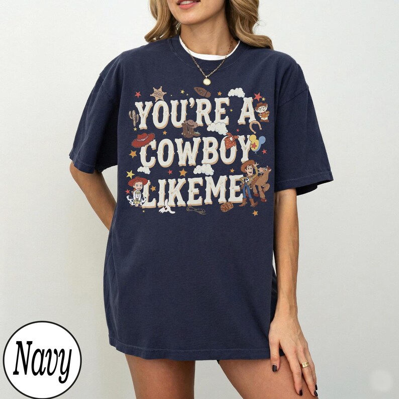 May include: A navy blue short-sleeve t-shirt featuring the text "YOU'RE A COWBOY LIKEME" in large cream-colored letters. The design includes small illustrations of cowboy hats, stars, balloons, and cartoon characters in a western theme.