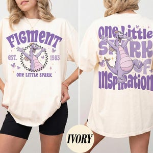 May include: Ivory-colored t-shirt featuring a purple Figment graphic with the text "Figment est. 1983" on the front. The back of the shirt displays a matching Figment graphic and the words "One Little Spark of Inspiration."