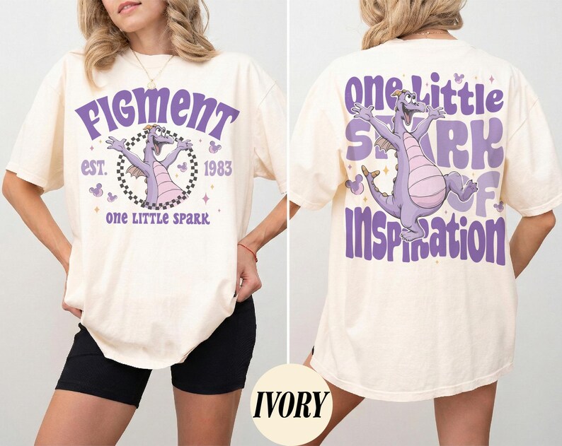 May include: Ivory-colored t-shirt featuring a purple Figment graphic with the text "Figment est. 1983" on the front. The back of the shirt displays a matching Figment graphic and the words "One Little Spark of Inspiration."