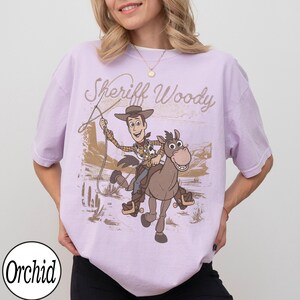May include: A light purple t-shirt featuring a graphic of Sheriff Woody riding a horse with the text "Sheriff Woody". The design is in shades of brown and tan, set against a desert backdrop. The shirt has short sleeves and a relaxed fit.